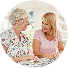 A middle aged woman sitting next to an elderly woman both happily working on a jigsaw puzzle