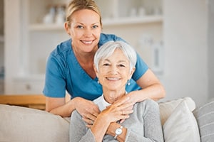 PACS-300x200-caregiver3 A caregiver wearing blue scrubs crouching over to an elderly woman on a sofa