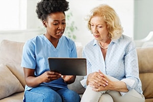PACS-300x200-computer2 A caregiver in blue scrubs showing a tablet computer to an elderly resident sitting on a sofa